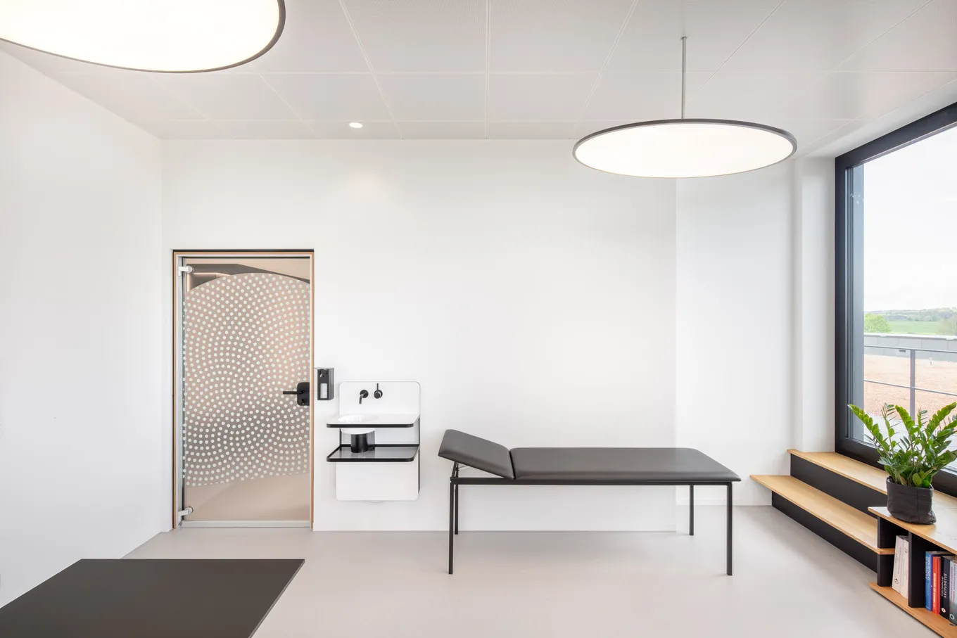 Modernes Behandlungszimmer mit großen Fenstern und einer medizinischen Liege auf grauem Betonboden