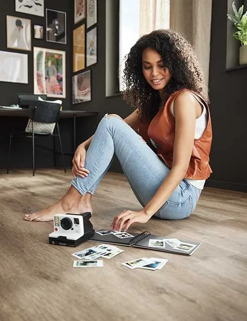 Frau sitzt mit mit Polaroidkamera und ausgedruckten Fotos auf einen Vinylboden i Holzoptik